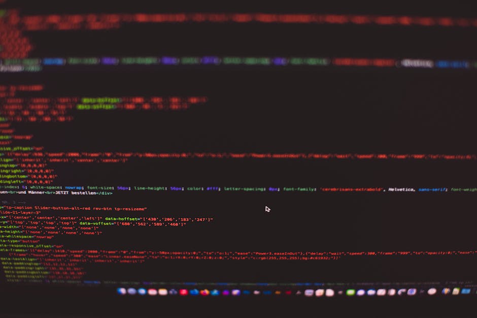 A person working on a MacBook laptop with a dark-themed coding interface displayed on the screen, featuring lines of colorful code and text editor windows. The individual's hands are visible, with one